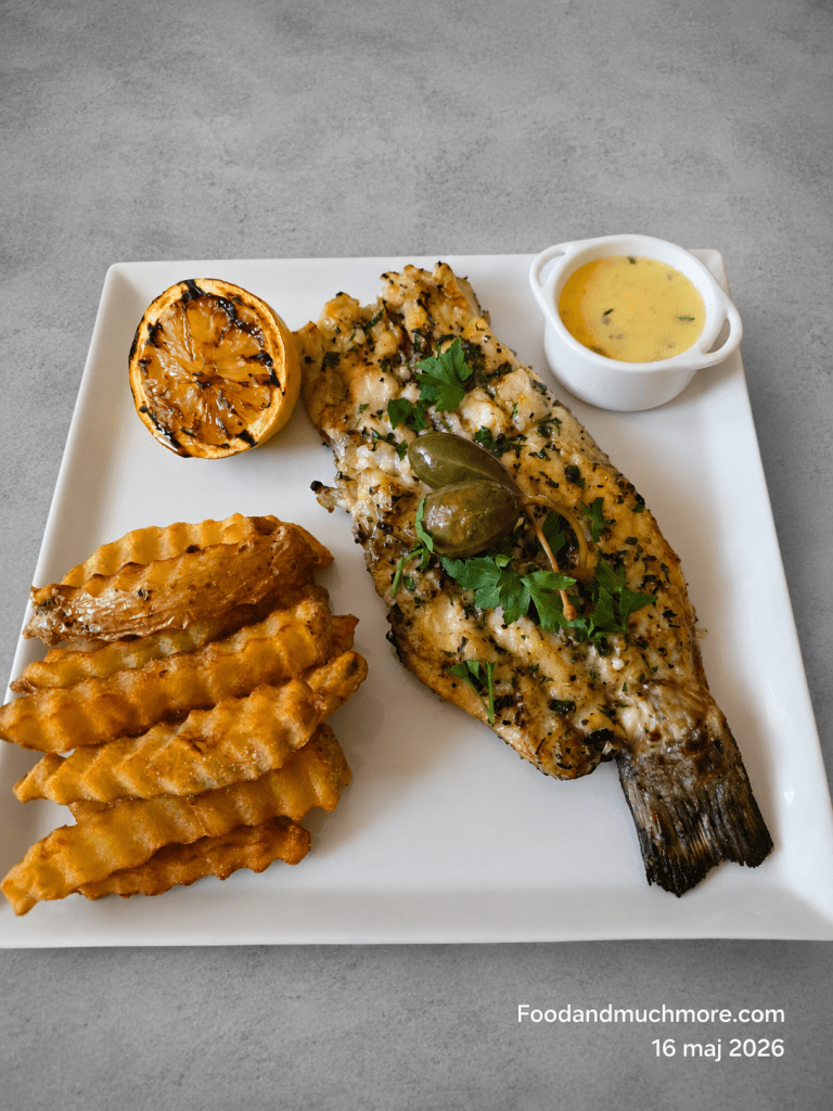 Grillad sea bass med hemmagjorda pommes, grillad citron och Beurre Blanc på vit tallrik mot grå bakgrund.